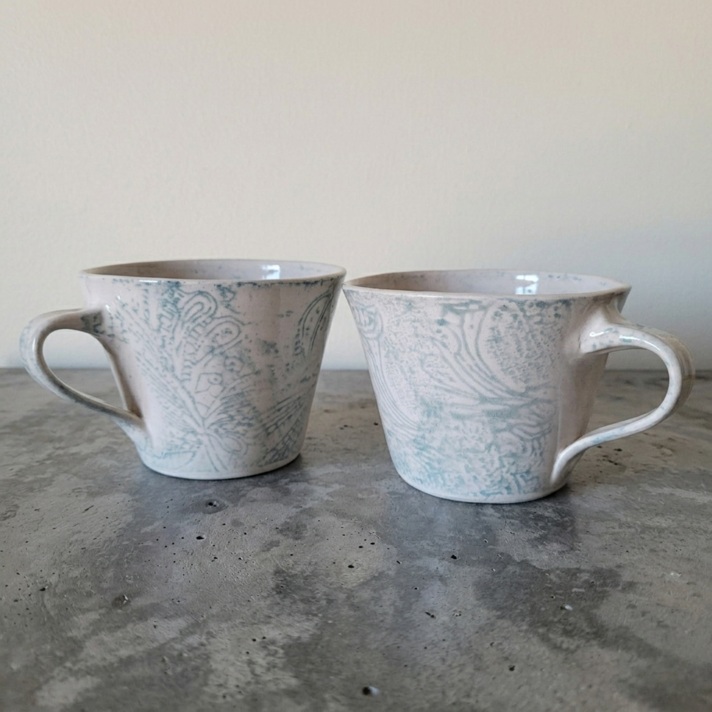 Two ceramic mugs with a paisley pattern on a concrete surface.