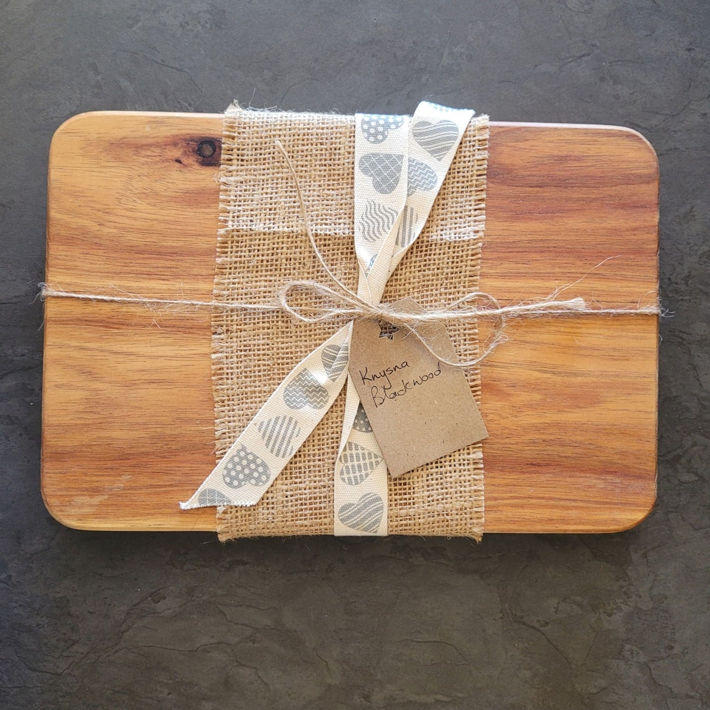 Wooden cutting board wrapped in burlap with a decorative bow on a dark surface