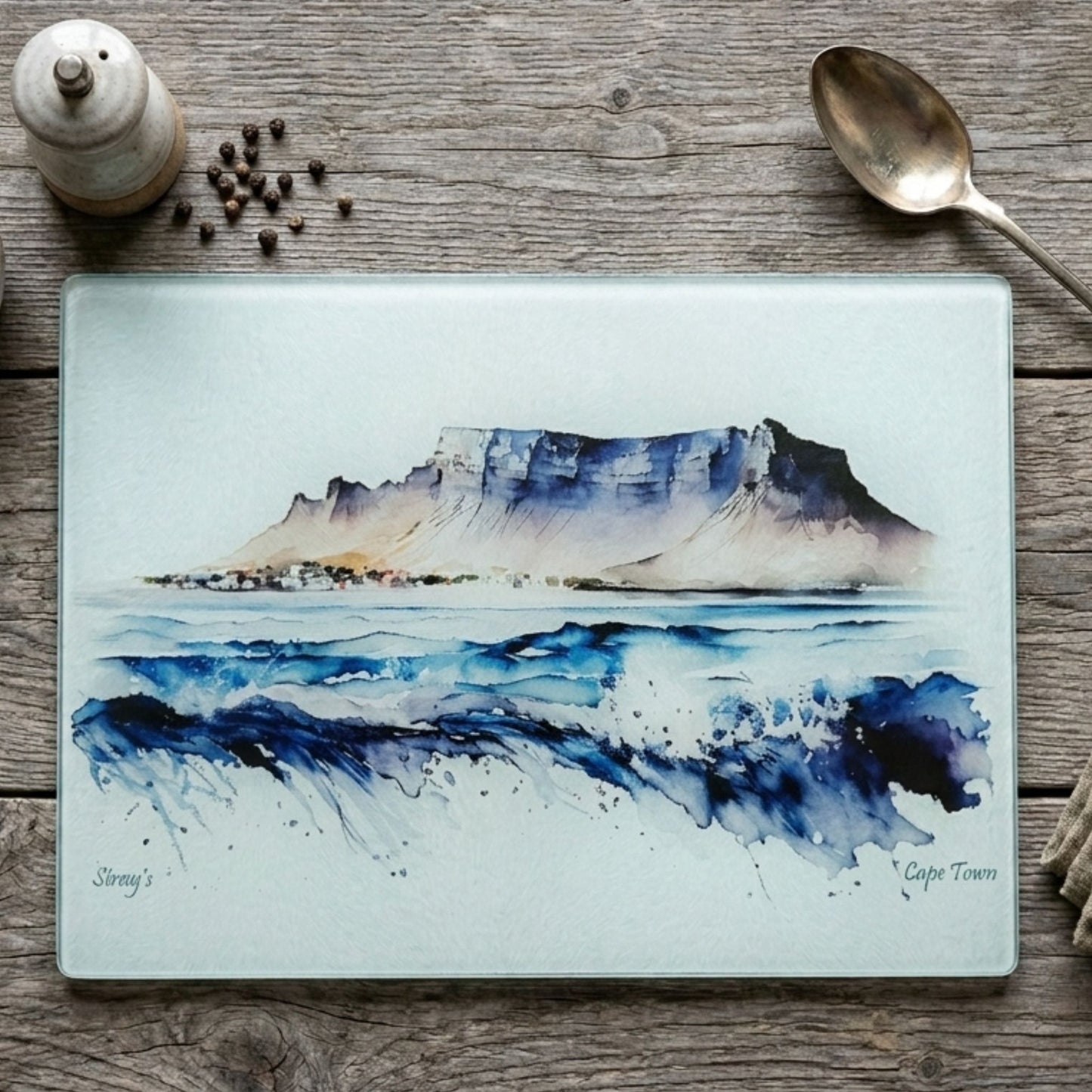 Glass cutting board with a mountain landscape on a wooden surface with a spoon and container.