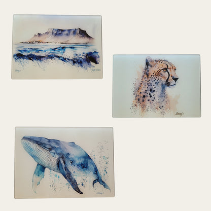 Three artistic illustrations of a mountain, cheetah, and whale glass cutting boards on a white background.