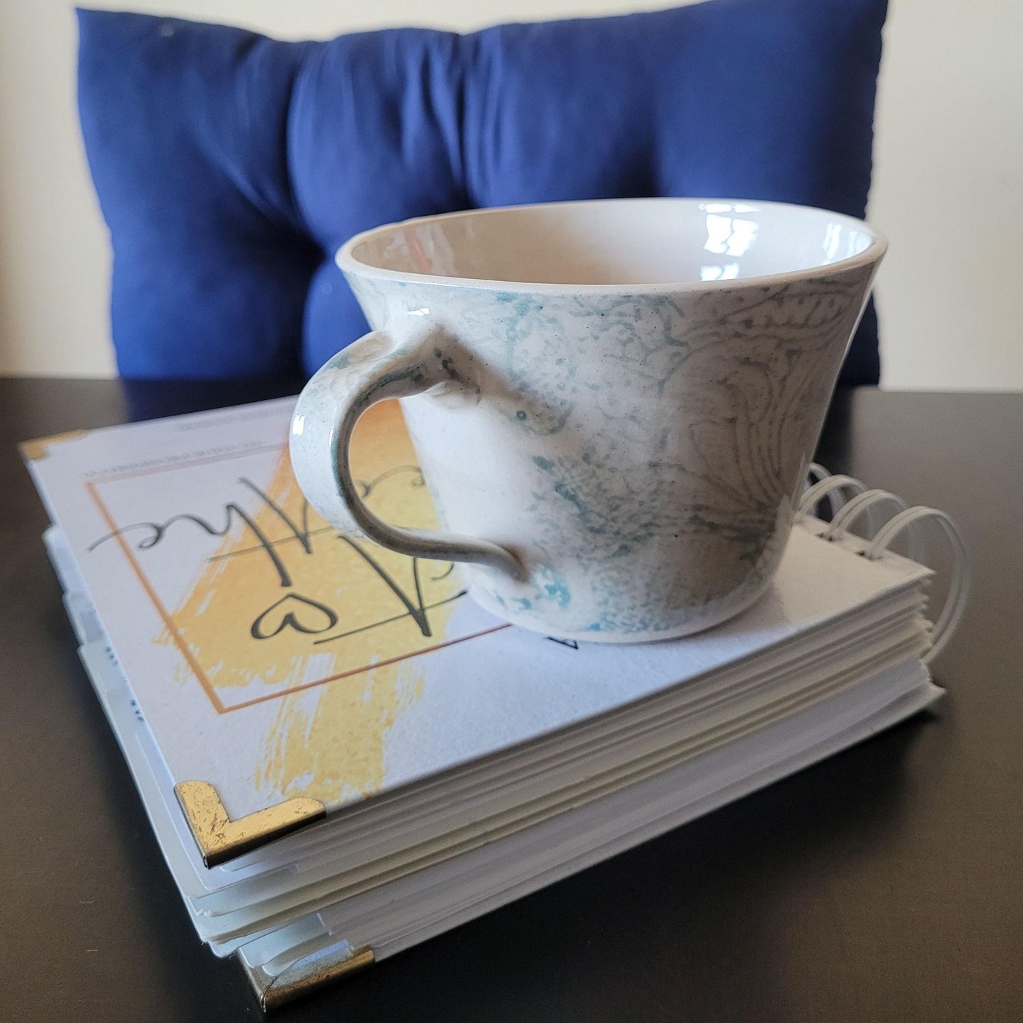 Patterned ceramic teacup with decorative patterns on a stack of books, with a blue cushion in the background.
