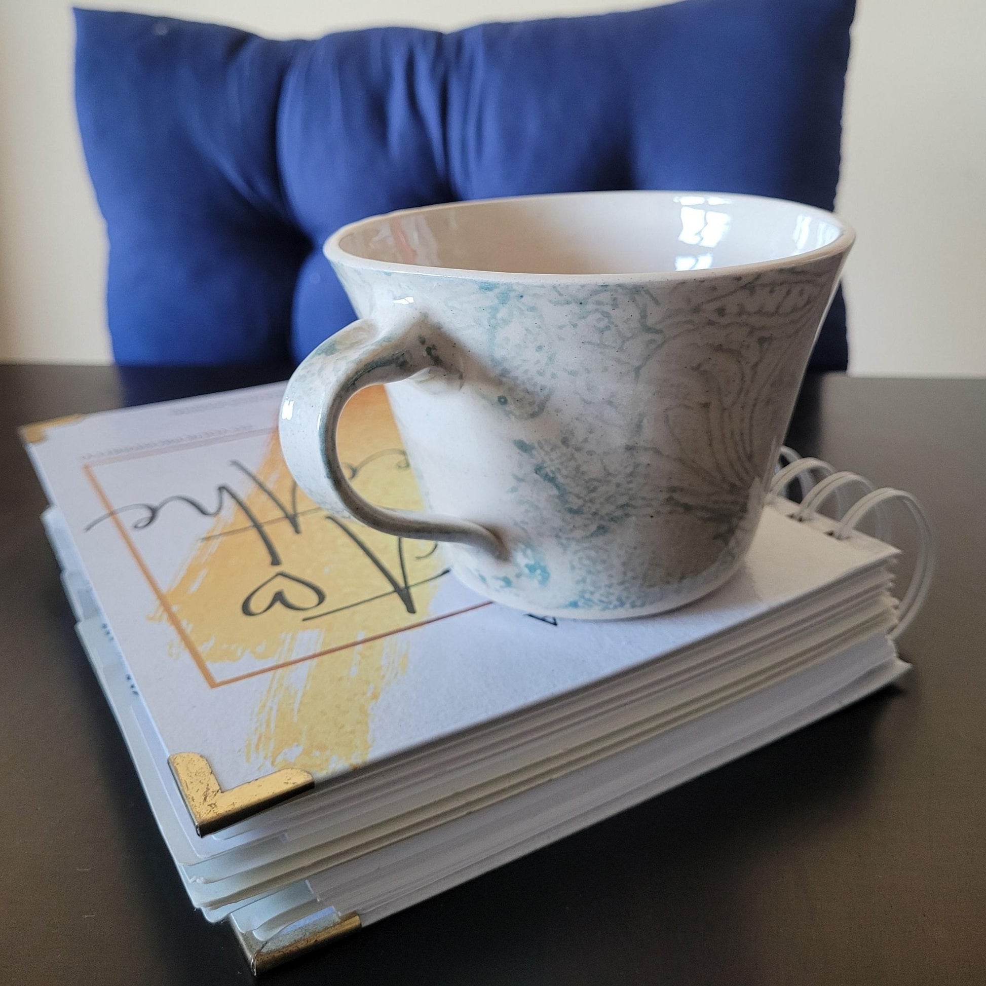Patterned ceramic teacup with decorative patterns on a stack of books, with a blue cushion in the background.